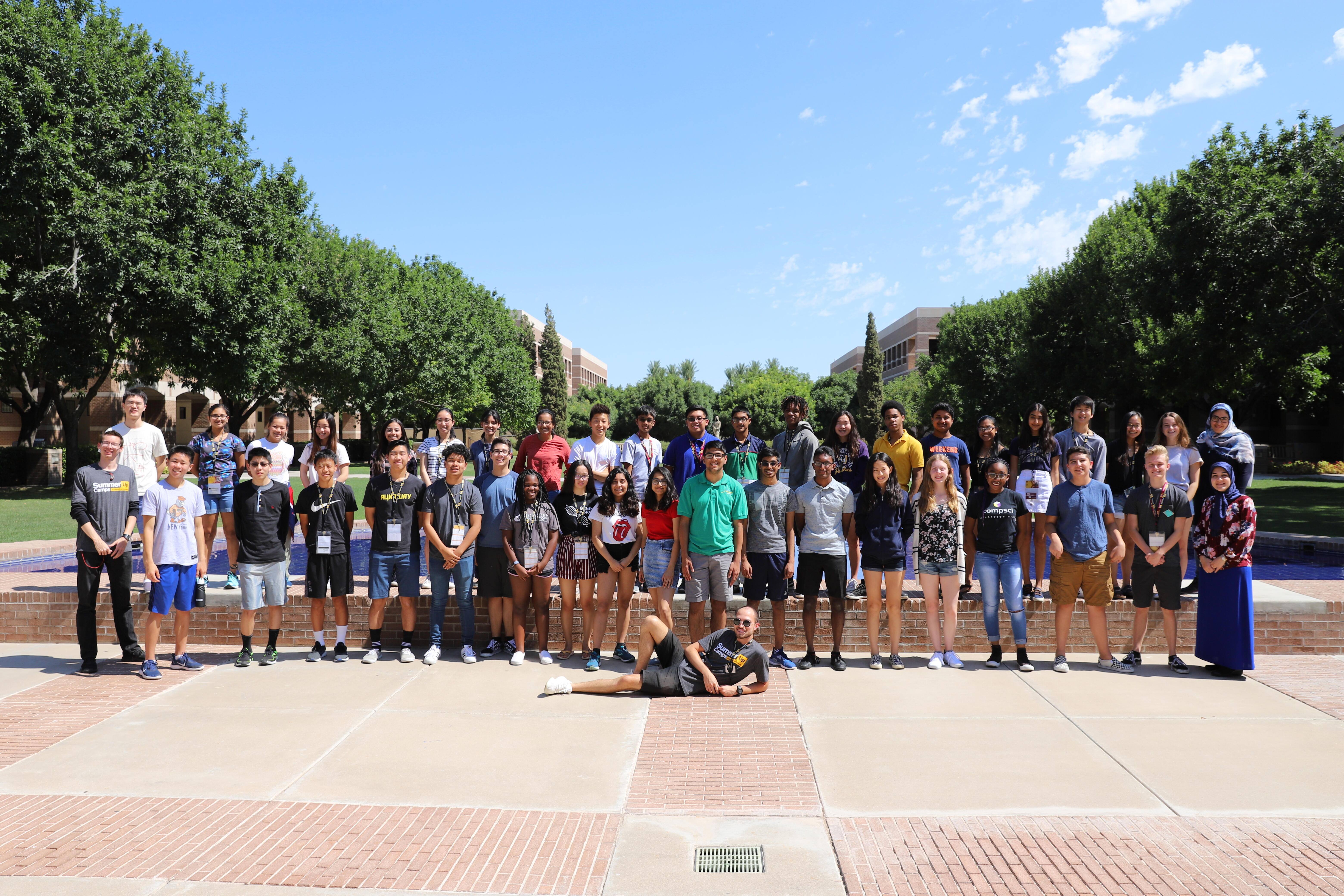 A group of 31 people posing outdoors in a courtyard with trees and buildings in the background on a sunny day.