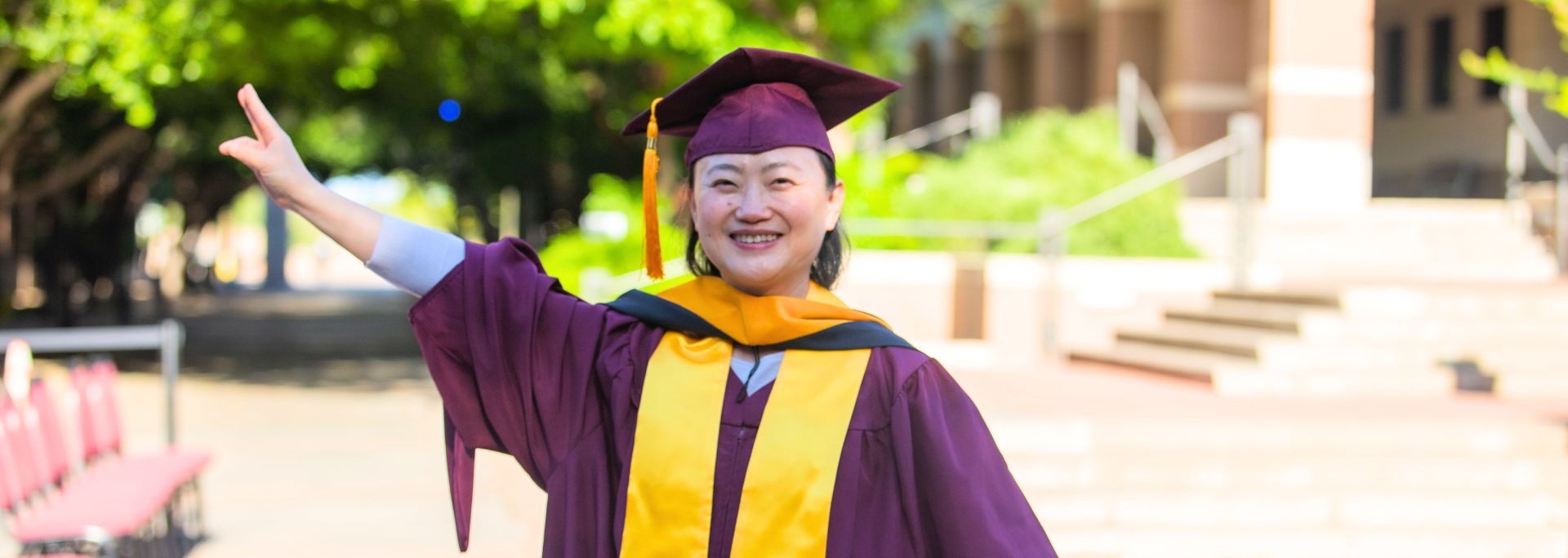 Jun Qian smiling outside while holding up an ASU pitch fork and wearing regalia