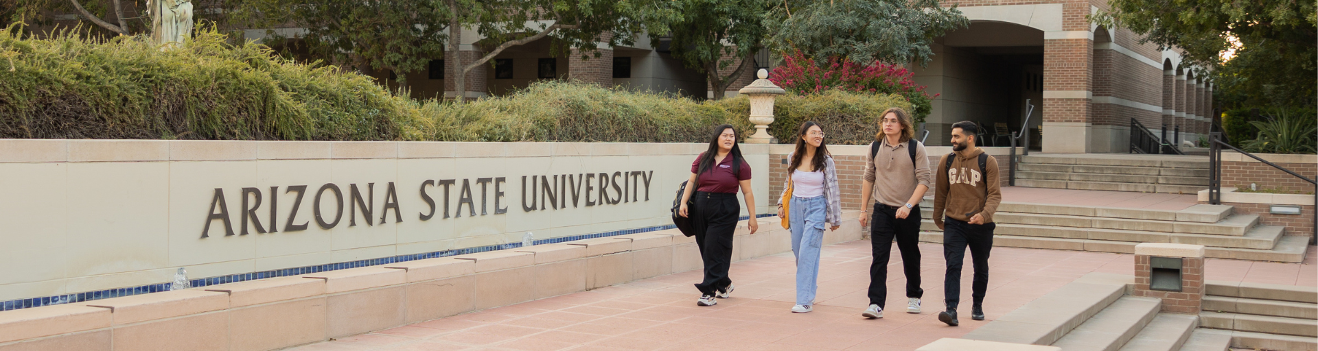People walking in front of an Arizona State University wall with greenery and a building in the background.