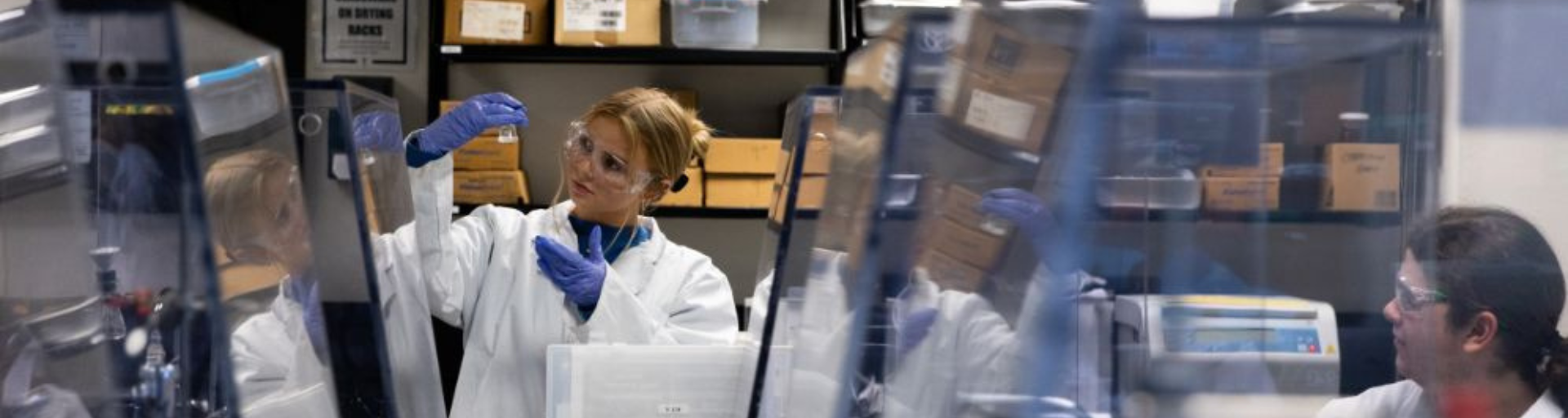 A scientist examines a vial in a laboratory surrounded by shelves with boxes.