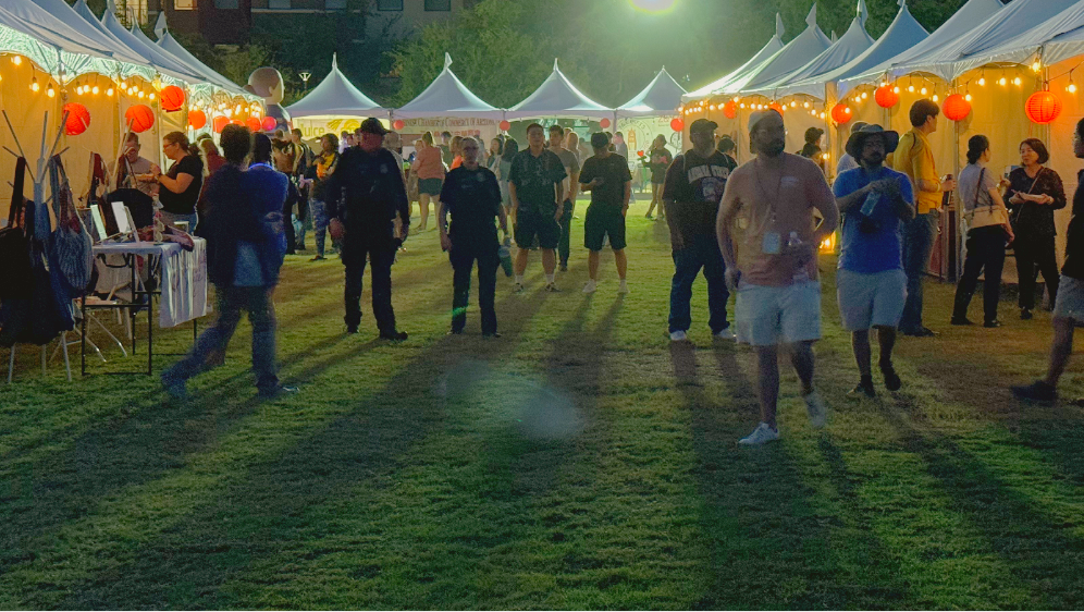 Night market scene with illuminated tents, red lanterns, and people walking on grass.
