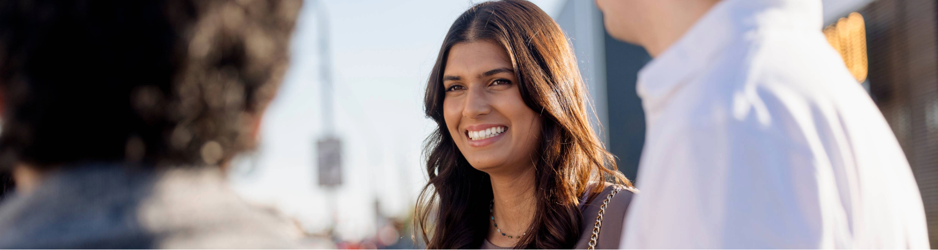A smiling woman with long dark hair and a necklace, outdoors with two partially visible people.