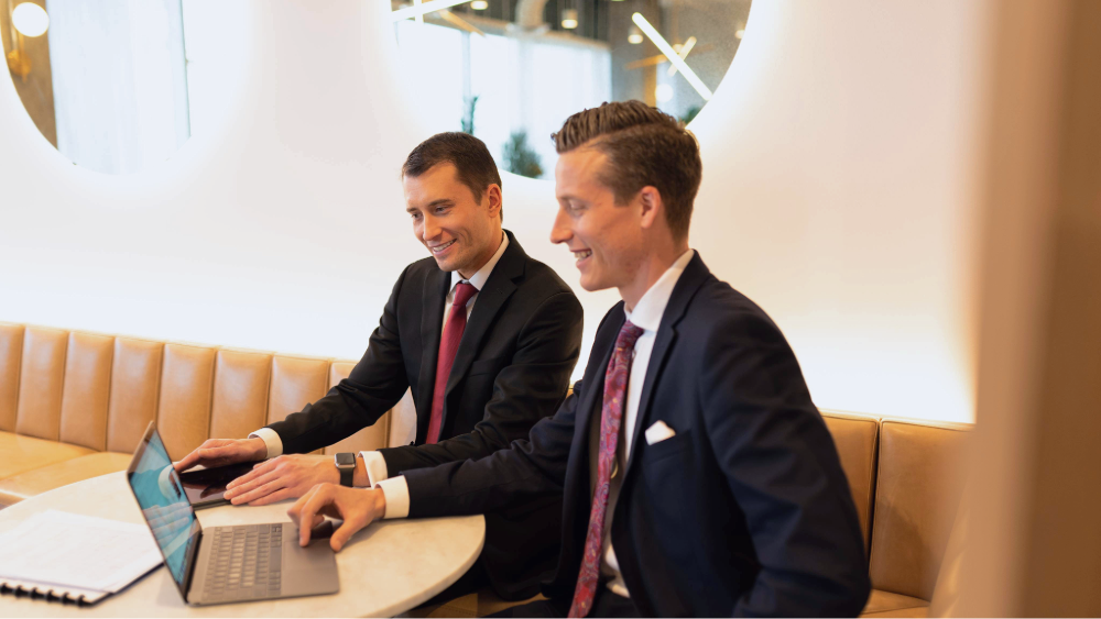 Two men in business suits smiling and interacting with a laptop at a round table.