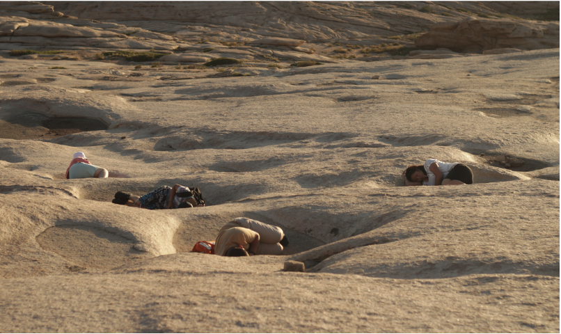 People lying face down in depressions on a sandy, rocky landscape during late afternoon.